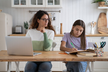 Work-at-home parent mother looking at daughter doing homework, sit together at table in kitchen. School girl studying with mom. Working online while kids. Parenting and remote work, homeschooling