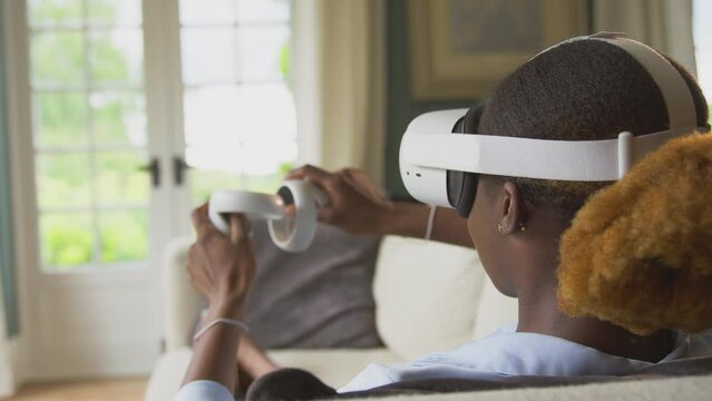 Woman lying on sofa at home wearing VR headset with controllers and interacting with AR technology - shot in slow motion
