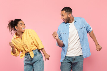 Joyful african american couple in casual posing together on pink studio background, dancing and smiling at each other