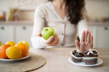 No junk food. Woman refusing to eat sweet cupcakes choosing apple, sitting in kitchen interior at home, selective focus