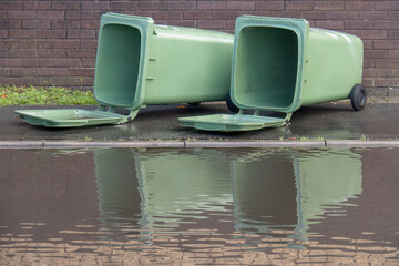 Two large green rubbish bins, lying in UK street, with large puddle, floodwater. After storm