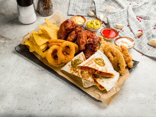assorted beer snacks, chicken wings, chicken strips, chicken quesadilla, onion rings, nachos on grey table