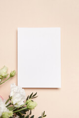 Holiday greeting card mockup with flowers on light beige background, top view, flat lay. White wedding invitation card mockup and floral decor