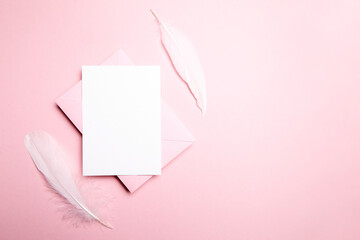 Holiday greeting card mockup with pink envelope and white feather on light pink background, top...