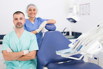 Handsome dentist with young female assistant in uniform with crossed hands at the dental office