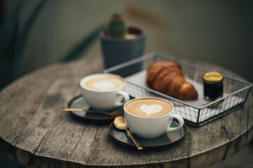 Two cups of cappuccino and fresh croissant with jam on wooden table.