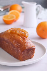 A whole orange cake topped with its sliced candied in a white plate with oranges fruit, plate and mug in background, it's a moist and soft sweet cake with entire orange used