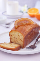 Close up a whole orange cake topped with its sliced candied in a white plate with its fruits with fork. mug and tableware in background, it's a moist and soft sweet cake with entire orange used