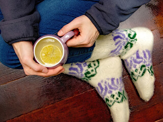 Female hands with a cup of hot tea and female legs in warm knitted socks on a dark wooden background