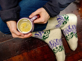 Female hands with a cup of hot tea and female legs in warm knitted socks on a dark wooden background
