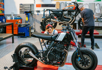 Mechanic female checking custom motorcycle while her colleague adjusting motorbike wheel over platform on garage