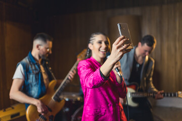 a happy singer wearing a pink blazer taking a selfie with a microphone and her band in the background, music studio. High quality photo