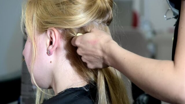 Hand of hairdresser combs through long strand of blonde lady. Female stylist creates curls twisting hair on modern styler in beauty salon closeup backside view