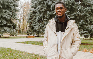 Cheerful young black man in stylish clothes walking in autumn park. Happy smiling guy in warm casual clothes over pine trees background