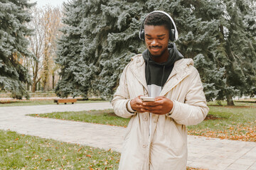 Cheerful black man in headphones is typing message on smartphone over park background. Smiling African guy in casual clothes and in white earphones walking on autumn square