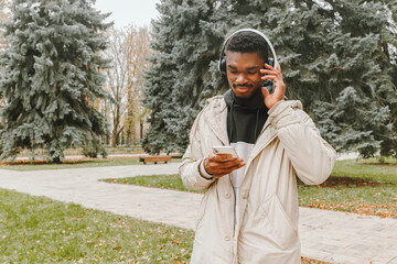 Handsome black man in headphones searching songs in playlist over park background. Smiling guy in warm casual clothes with wireless earphones walking on autumn day