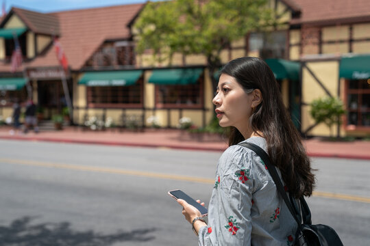 confused asian japanese female traveler looking into space to check directions while using navigation app on the phone on the main street of solvang California usa