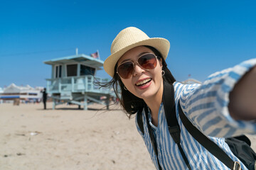 happy asian Taiwanese female traveler wearing hat and sunglasses having fun taking selfie photo on santa monica beach with lifeguard tower in back.