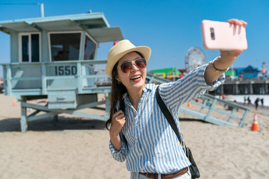 happy asian Korean woman traveler having fun using smartphone to take photo of herself with famous Lifeguard Tower at background at santa monica beach on sunny day