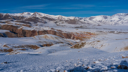 Picturesque unusual mountains with red-brown slopes against a blue sky. In the foreground is an untouched snow-covered plateau. The dry grass is covered with hoarfrost.  Altai Mars Kyzyl Chin