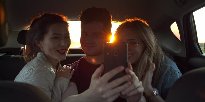 Fun Young Womens and man laugh and do a selfie In Back Seat Of Moving Car. Slow Motion. Sunset on background. flares