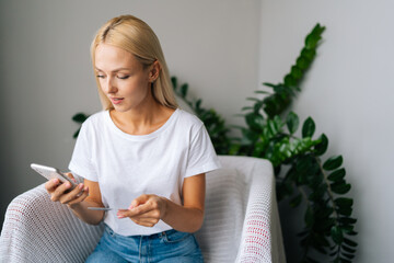 Portrait of focused young woman sit on bed at light bedroom holding plastic credit card, provide...