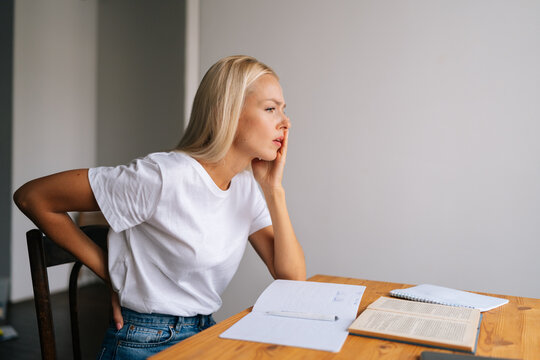 Side view of female student sit at desk touch back suffer from lower spinal spasm, hurt unhealthy biracial woman stretch have strong backache, sitting at desk with book and notebook at home.