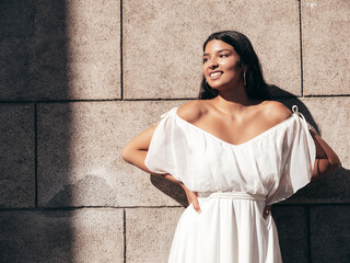 Young beautiful smiling hipster woman in white dress. Sexy carefree model posing on the street background at sunset. Positive female outdoors at warm sunny day. Cheerful and happy