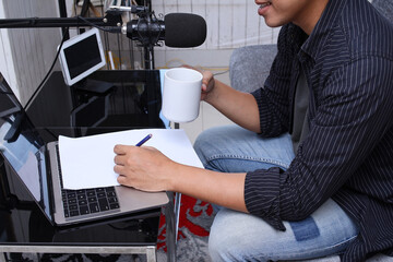 Asian man recording a podcast on his laptop while writing on blank paper at home studio