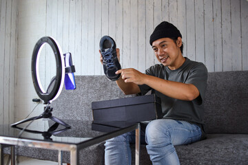 Smiling Asian male influencer or shoes seller showing his product to online followers through smartphone. Product review video