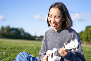 Happy people and hobbies. Smiling asian girl playing ukulele guitar and singing, sitting in park...