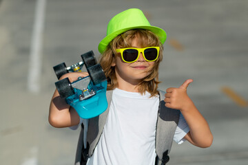 Amazed child with thumb up, street portrait. Little kid boy 7-8 years old hold skateboard posing on street outdoor. Childhood lifestyle concept. Children skateboarding.