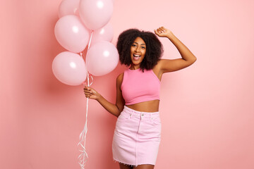 Positive cheerful young woman with curly hair dances joyfully with balloons in a hand, keeps arms raised up, dressed in pink top and skirt, isolated over pink background