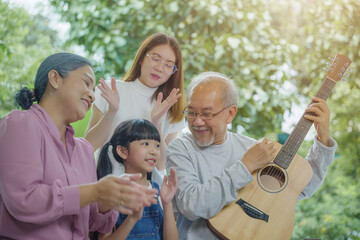 Happy family Asian senior elderly man or grandfather playing guitar while his grandmother and granddaughter singing song together outdoors at home, Activity family, Enjoying lifestyle senior older