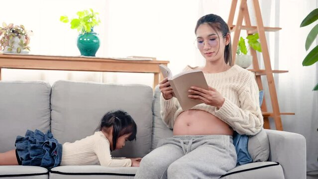 4K, Pregnant Asian single mom reads a book on sofa in her bedroom with her adorable daughter lying on her mobile phone next, Out of concern and love for her child, she often turned to see her child.