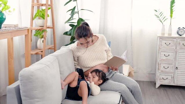 4K, Pregnant Asian single mom reads book on sofa in her bedroom with Lovely daughter laying down playing mobile phone games on lap do not have fun, two of them talked, taking care of child with love.