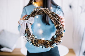 Very nice young woman holding big and beautiful hand made Christmas festive wreath decorated with dry limes, pine cones, acorns, bow in light blue colors cropped photo, bouquet close up
