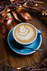 Ceramic blue cup of delicious fresh cappuccino coffee on the rustic wooden background decorated with festive hand made wreath, fireflies and copper painter leaves