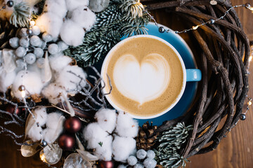 Delicious fresh cup of cappuccino coffee standing in hand made wooden wreath decorated with fireflies, cotton, pine cones, spruce branches, berries on the wooden table background, top view, flat lay