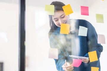 Portrait shot of Cheerful young adult business Asian woman sticking adhesive paper notes to a glass wall, idea-sharing board in business office concept.