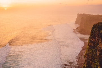 Ocean, waves and cliff at warm sunset with fog in Uluwatu, Bali