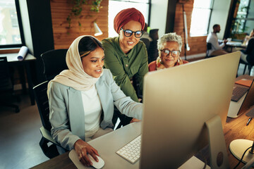 Diverse group of female coworkers use a computer together in a creative, online workplace, smiling and working collaboratively as they pursue their business goals.