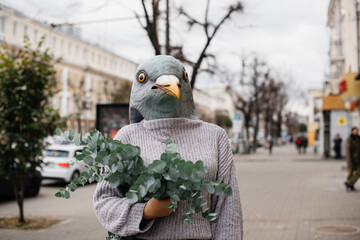 Weird woman a bird (pigeon) mask holding a bunch of fresh eucalyptus, standing in the street - 555058706