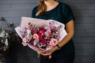 Very nice young woman holding big and beautiful bouquet of fresh delphinium, roses, eustoma, eucalyptus in pink and purple colors, on the dark grey wall background, bouquet close up