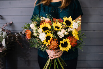 Very nice young woman holding big and beautiful bouquet of fresh nuts, sunflowers, roses, eustoma, pistachio in yellow and orange colors, on the dark grey wall background - 555059123