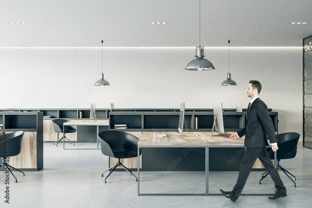 Poster Businessman in black suit walking through wooden work table with modern computers in open space modern office with dark stylish furniture and light wall background