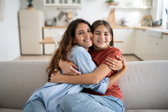 Young woman loving mother hugging embracing her happy adopted daughter at home, expressing love to teenage child, joyful mom and child sitting together on couch and cuddling. Family reunion concept