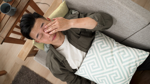 View From Above Of A Sleepy Asian Young Man Lying On Sofa After Yawning Rubbing Eyes At Home.