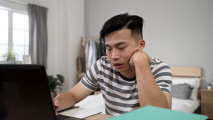 sleepy asian male student learning from home is propping face and nodding off while taking online...