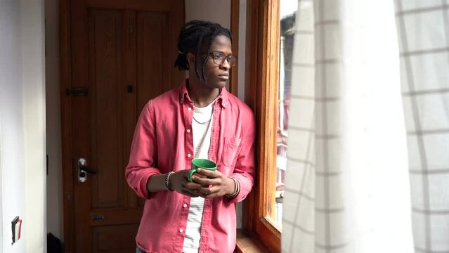 Young sad depressed African guy crying while standing with cup of tea by window at home. Upset emotional black man dealing with grief, coping with loss of loved one. Depression after bereavement